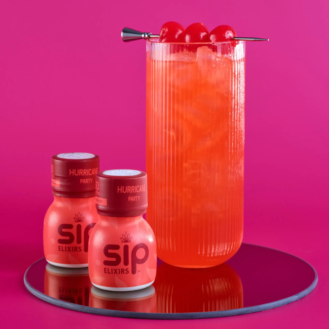 Red cocktail with cherries and 'sip Elixirs' bottles on a pink background