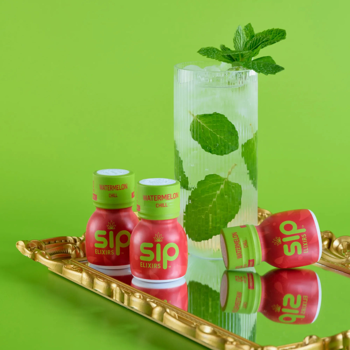 Two bottles of Sip Elixirs watermelon chill on a gold tray with a glass of green drink and mint leaves against a green background.