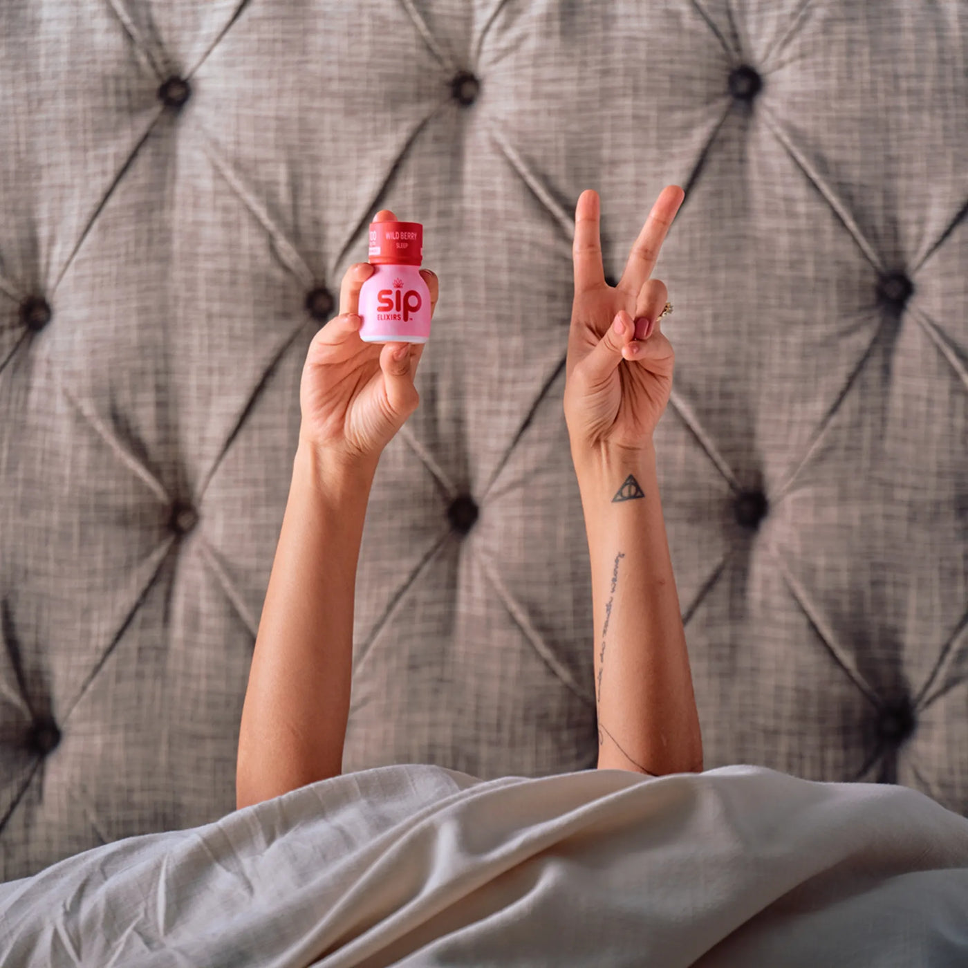 Person lying in bed holding a pink bottle with 'sip' branding, making a peace sign.